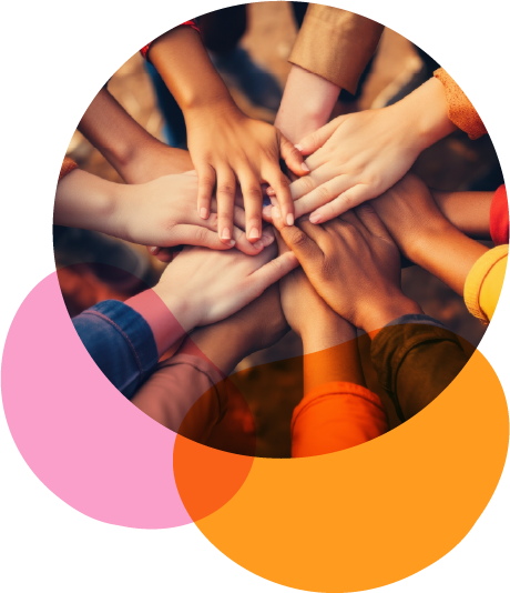 Grupo de pessoas em pé formando um círculo. No centro, todas estendem as mãos umas sobre as outras, empilhando-as em sinal de união. As mãos são de diferentes tons de pele, representando diversidade. As mangas das roupas são coloridas — há tons de vermelho, amarelo, laranja, marrom e azul. O gesto transmite sentimento de acolhimento, solidariedade e trabalho em equipe. O chão é de terra, sugerindo que estão ao ar livre.